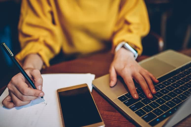 Female entrepreneur sitting on her desk, writing on her notebook with her laptop.