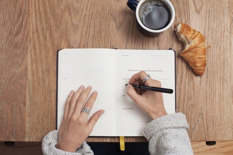 A woman writes in a notebook on a table with a cup of coffee and a croissant.
